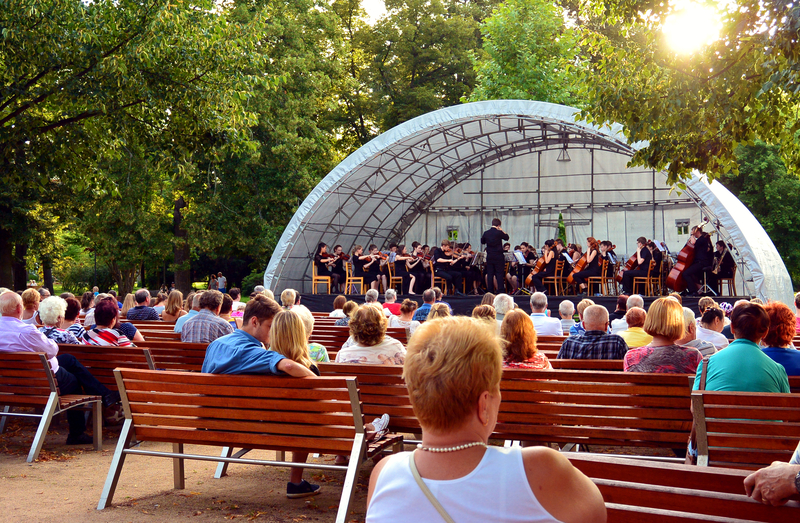 A group of rural community members attend a concert in a park. The stage is covered in a partial dome.