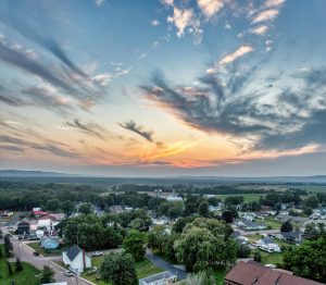 Landscape of a rural town pockmarked with trees beneath a dramatic sky with a pastel orange sunset along the horizon.